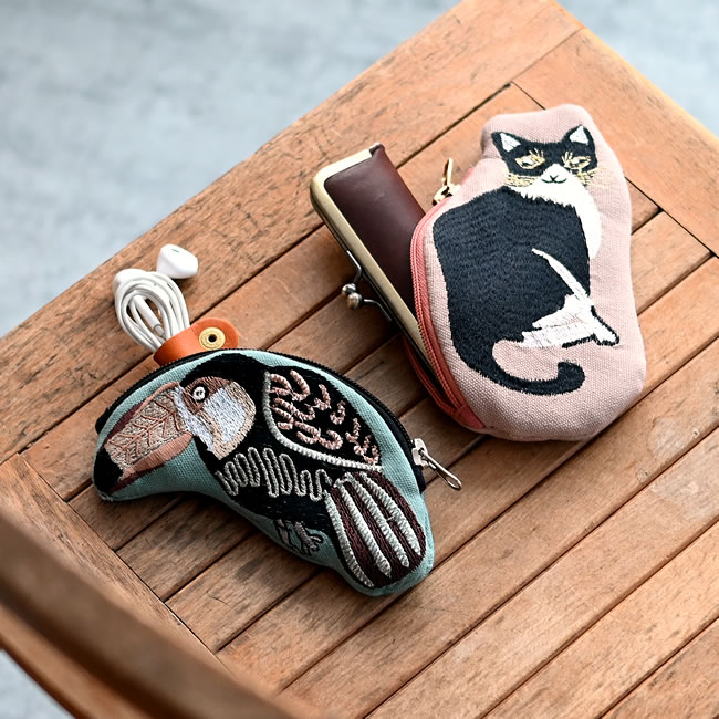 イヤホンや小物、がま口と一緒に置かれたオオハシと黒ネコの刺繍ポーチ
